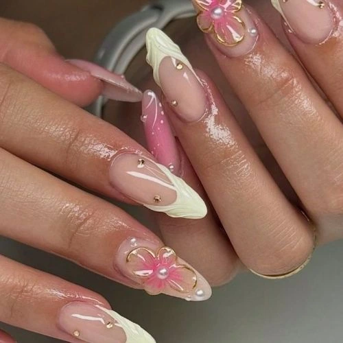 A set of long, almond-shaped nails with a soft pink base, adorned with delicate 3D floral designs, gold embellishments, and creamy white accents.