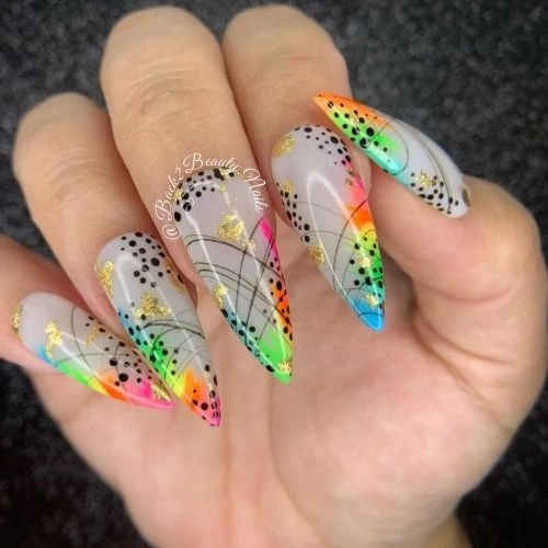 A hand with long, pointed nails showcasing a spring-inspired design. The nails have a light gray base color with vibrant neon accents in shades of green, yellow, orange, pink, and blue. The design is enhanced with black dots, gold foil details, and fine black lines forming an abstract pattern.