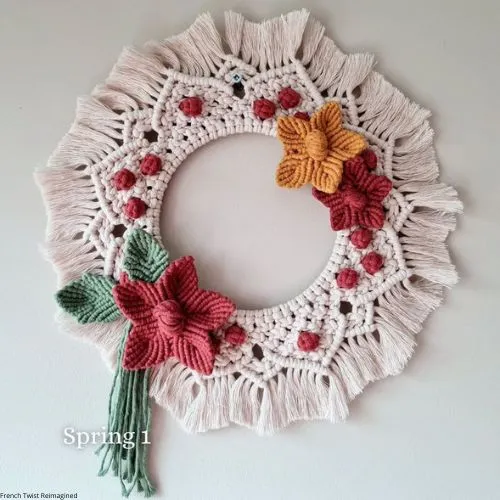 A handcrafted macramé wreath with intricate patterns, featuring crocheted red and orange flowers and tassel fringe.