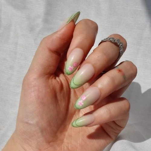Hand with almond-shaped nails featuring a sheer nude base, soft green French tips, and tiny hand-painted pink flowers, photographed in natural sunlight.