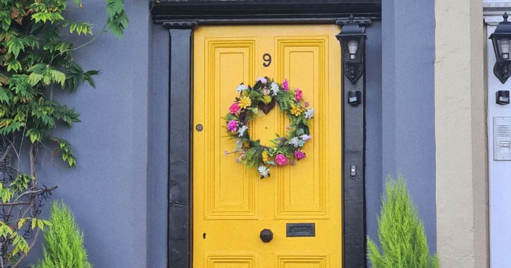 A vibrant floral wreath with pink, yellow, and white flowers hanging on a bright yellow door with a black frame.