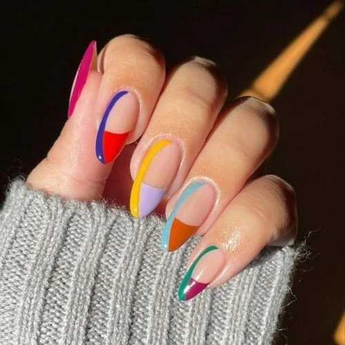 A close-up image of a hand with colorful spring-themed nail art. Each nail displays a unique design using vibrant hues such as red, blue, yellow, purple, teal, and orange. The hand is wrapped in a gray knitted sweater.