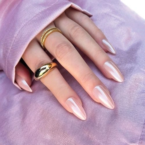 A close-up of a hand with glossy, almond-shaped nails painted in a soft pink hue. The hand is partially wrapped in a silky, light pink sleeve, exuding a spring-inspired elegance. The nails are paired with two gold rings—one a chunky statement piece and the other a delicate band with small diamonds.