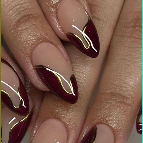 A set of almond-shaped nails with a nude base and deep burgundy French tips, accented with sleek gold lines in a modern negative space design.