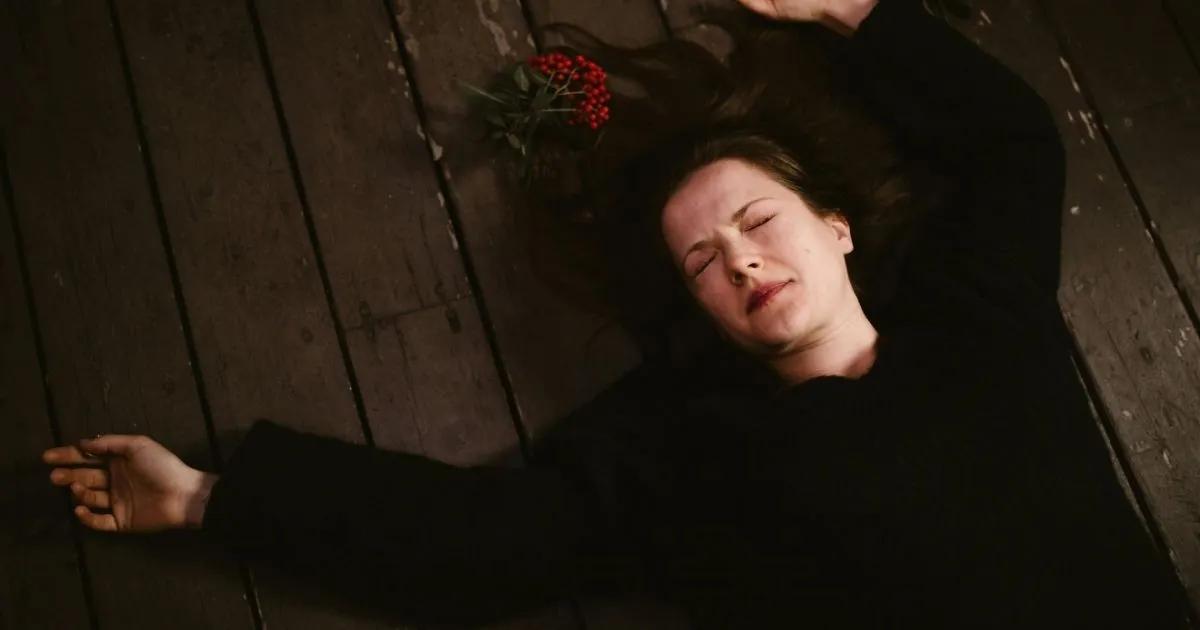 A woman in a black sweater lying on a dark wooden floor with her arms spread out and her eyes closed, with red berries near her head. red flags in men