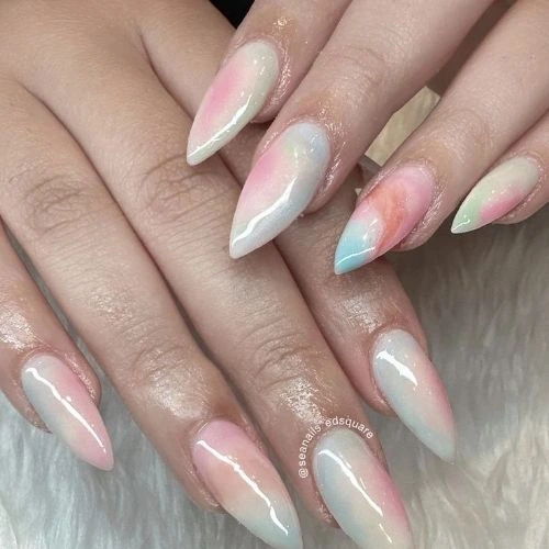 Close-up of hands with almond-shaped nails featuring a pastel watercolor ombré effect in pink, blue, and peach, finished with a glossy sheen.