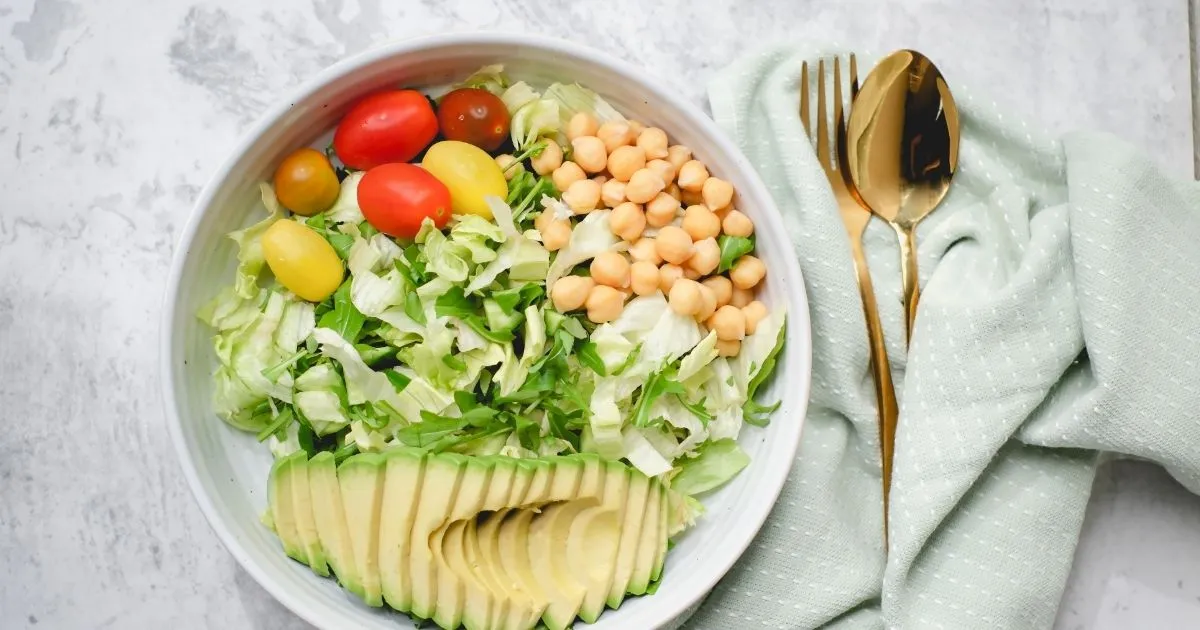 A white bowl containing a balanced salad with greens, chickpeas, cherry tomatoes, and sliced avocado.