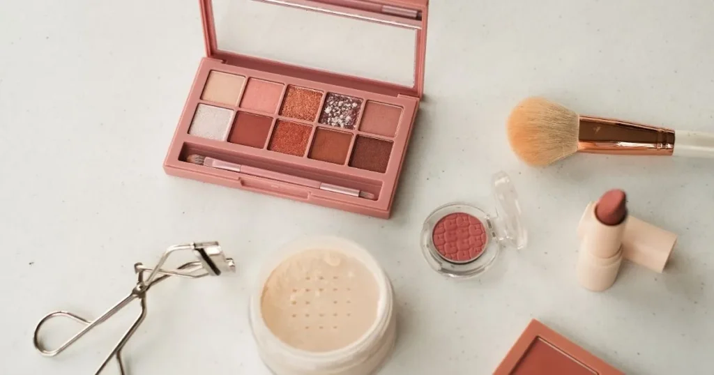 An artistic display of makeup essentials, including an eyeshadow palette, a makeup brush, a lipstick, and a blush compact, arranged on a flat surface.