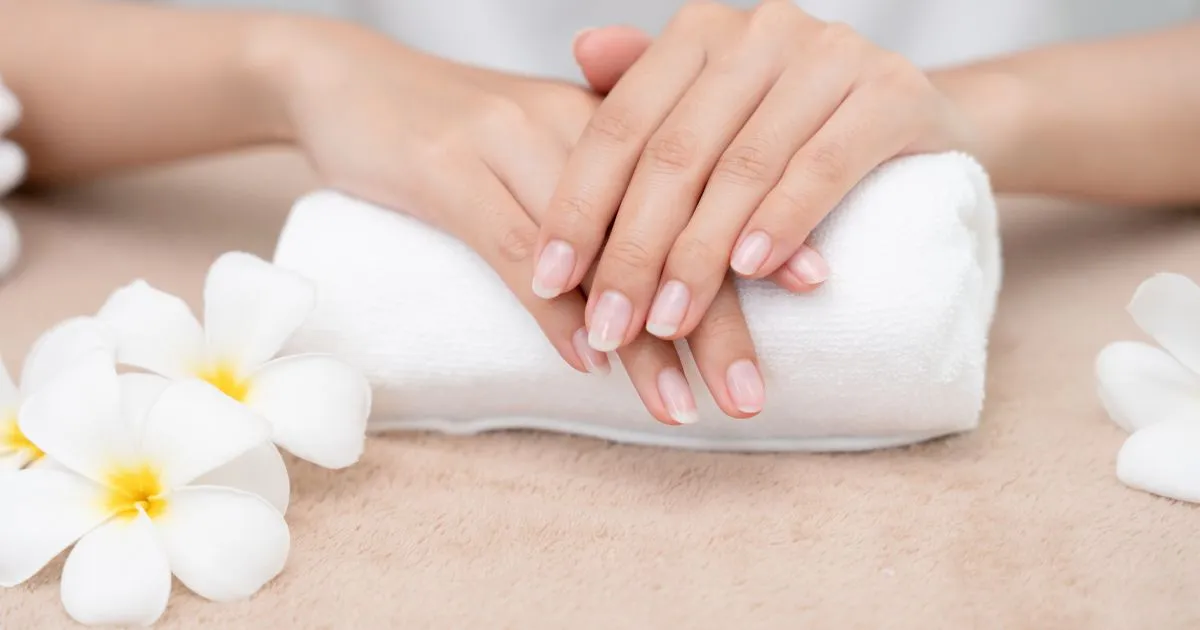 A serene spa scene featuring a pair of hands resting on a rolled white towel, surrounded by delicate white frangipani flowers with yellow centers. The hands showcase natural, clean nails, contributing to the calm and elegant atmosphere #Spring_Nail